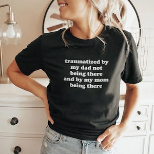 Puede incluir: Camiseta negra de manga corta con texto blanco: "traumatized by my dad not being there and by my mom being there." La camiseta la lleva una persona. La persona lleva vaqueros azules.