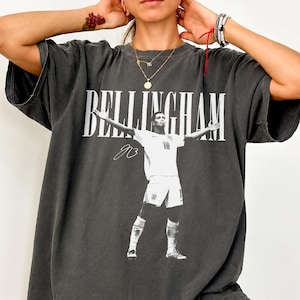 May include: A charcoal grey short-sleeved t-shirt featuring a white graphic print of a football player with arms outstretched. The text BELLINGHAM is printed in large white capital letters above the player, with a signature graphic below the name.