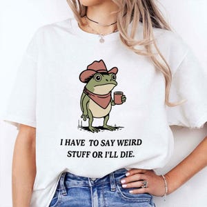 Puede incluir: Camiseta blanca con una rana de dibujos animados con sombrero de vaquero y taza. La rana es verde, marrón y beige, con un pañuelo rojo. El texto "I HAVE TO SAY WEIRD STUFF OR I'LL DIE" está impreso debajo.