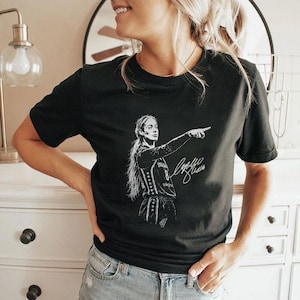 May include: A black short-sleeved t-shirt featuring a white graphic illustration of a basketball player pointing with one arm extended. The design includes the player wearing a jersey with the number 8 and a stylized signature written in white ink.