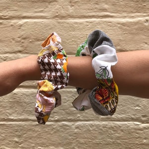Puede incluir: Dos scrunchies de tela, uno con un patrón de pata de gallo marrón y blanco y un estampado floral, y el otro con un fondo blanco y un estampado floral con un patrón de pata de gallo negro y blanco.