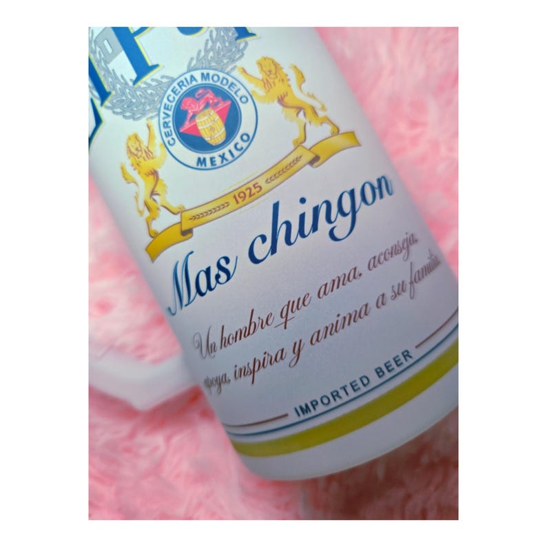 Peut inclure: Gros plan sur une canette de bi&egrave;re avec le texte "Mas chingon" en &eacute;criture bleue. La canette pr&eacute;sente un embl&egrave;me or et bleu avec les mots "Cerveceria Modelo Mexico" et "Imported Beer". Le fond est rose.