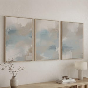 Pode incluir: Um conjunto de três peças de arte de parede retangulares em molduras de madeira clara, com padrões de pinceladas abstratas em tons suaves de azul, bege e branco, dispostos horizontalmente para criar uma exibição visual coesa na parede.
