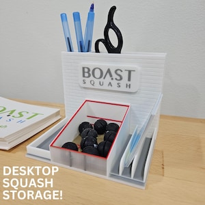 Op de afbeelding: Witte bureau-organizer met de tekst "BOAST SQUASH" en "DESKTOP SQUASH STORAGE!". Het heeft compartimenten voor pennen, scharen, visitekaartjes en squashballen. De organizer staat op een houten oppervlak.