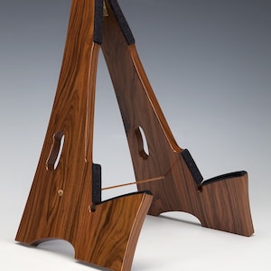 May include: A wooden A-frame guitar stand with a dark brown finish. The stand has a curved base and padded supports. The design includes cut-out details and a horizontal support rod. The stand is made of wood.