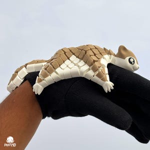 May include: A 3D printed articulated pangolin figurine featuring a segmented body with alternating tan and white scales. The creature has a smooth tan head with large black and white eyes, a small snout, and a flexible, multi-jointed tail structure.