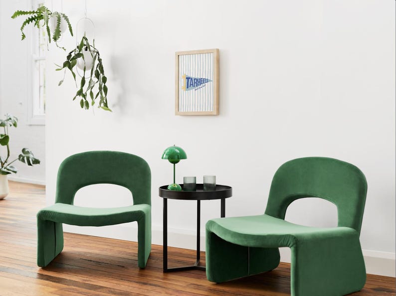 Peut inclure: Deux fauteuils en velours vert avec un dossier arrondi et une d&eacute;coupe. Une petite table d'appoint noire avec une lampe verte et deux verres se trouve entre les chaises. Une banni&egrave;re encadr&eacute;e avec le texte "TARHEELS" est accroch&eacute;e au mur blanc.