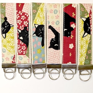 May include: Six fabric key fobs featuring black cat silhouettes on patterned backgrounds of red, green, and cream with floral and geometric motifs. Each strap is finished with a silver-toned metal clamp and a circular split ring for keys.