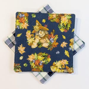 May include: A square quilted fabric coaster featuring a dark blue background with a repeating pattern of scarecrows and colourful autumn leaves in shades of yellow, orange, and green. A second coaster with a green and white plaid pattern is visible.