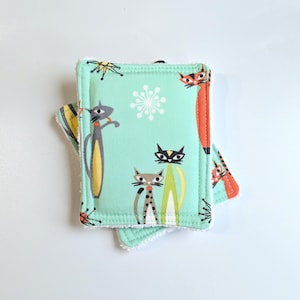 May include: Two turquoise blue dishcloths with a white border and a retro cat print. The cats are in various poses and colours, including orange, black, and grey. The dishcloths are made of a soft, absorbent fabric.