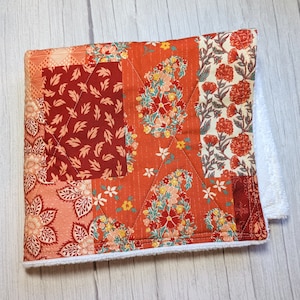 May include: A quilted fabric dishcloth with a patchwork design featuring various shades of red, orange, and white floral patterns. The dishcloth has a white terrycloth border.