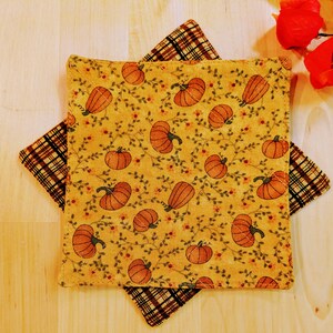 May include: Two square quilted pot holders. The top pot holder has a yellow background with orange pumpkins and green vines. The bottom pot holder has a brown and tan plaid pattern. The pot holders are likely for kitchen use.