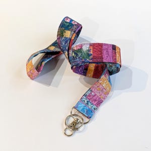 May include: A fabric lanyard featuring a vibrant patchwork design with various colourful patterns including shades of pink, blue, yellow, and purple. The strap is looped and includes a silver-toned metal swivel lobster clasp and a circular key ring.