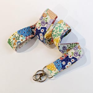 May include: A fabric lanyard featuring a colourful patchwork design with various floral, geometric, and plaid patterns in shades of blue, yellow, green, purple, and white. The strap is attached to a silver-toned metal swivel lobster claw clasp.