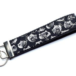 Può includere: Un portachiavi da polso nero con un motivo di facce di gatto bianche. Il tessuto presenta un motivo ripetuto di facce di gatti stilizzate. Dotato di anello e hardware in metallo argentato. Un accessorio pratico ed elegante.