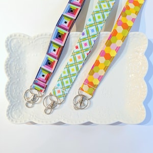 May include: Three fabric lanyards with metal swivel clasps rest on a white, scalloped-edge plate. The lanyards feature colorful patterns: a rainbow log cabin design, a light blue and green starburst pattern, and a yellow and pink honeycomb pattern.