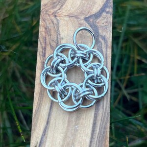 May include: A silver-colored pendant with a circular design made of interlocking rings. The pendant is attached to a small ring at the top. The background is a wooden surface with green grass.