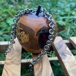 Könnte beinhalten: Ein holzfarbener, apfelförmiger Gegenstand mit dunklem Stiel, der ein natürliches Holzmaserungsmuster aufweist. Eine silberne Halskette liegt um den Apfel, der auf einer Holzoberfläche ruht.