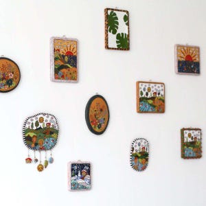 May include: A collection of colorful, framed art pieces with various designs, including landscapes, floral patterns, and a leaf motif. The frames vary in shape and style, creating a visually diverse wall display. The art pieces are mounted on a white wall.