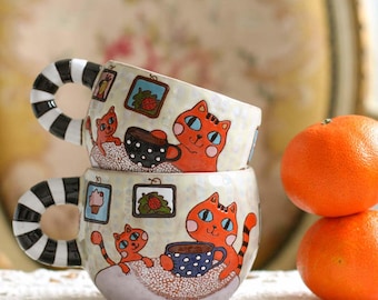 Puede incluir: Dos tazas de cerámica apiladas con una superficie texturizada de color crema. Cada taza presenta un gato naranja pintado a mano sosteniendo una taza azul de lunares, arte de pared enmarcado y un asa con rayas horizontales negras y blancas.