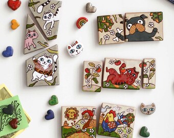 May include: A collection of ceramic tiles and small heart-shaped pieces featuring hand-painted illustrations of cats, a dog, a rooster, and a landscape with a house and sun. The tiles display colorful, whimsical scenes with floral motifs and patterns.