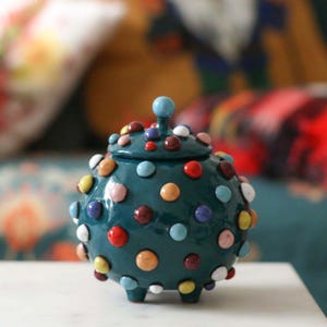 Op de afbeelding: Een teal keramische pot met een deksel, versierd met kleurrijke, verhoogde stippen. De pot heeft een kleine, ronde knop bovenop en drie kleine voetjes. De stippen zijn in verschillende kleuren, waaronder rood, geel, blauw en wit.