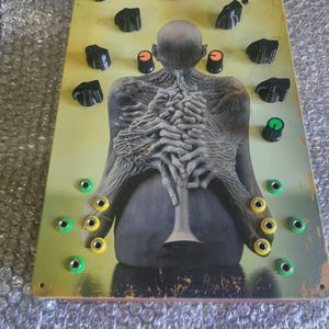 Op de afbeelding: Een rechthoekige elektronische synthesizer-module met een geel en bruin verloop op de voorplaat, voorzien van een centrale illustratie van een kaal figuur dat een trompet vasthoudt. Het apparaat bevat meerdere zwarte bedieningsknoppen, één roze knop en diverse groene en gele ingangen.