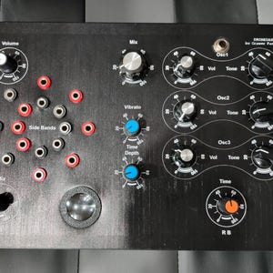 Puede incluir: Sintetizador electrónico negro con múltiples perillas, diales y conectores de entrada. Cuenta con controles etiquetados como "Volume", "Mix", "Vibrato", "Time" y "Side Bands". El texto "DRONESAX by Grawer Pasieka" está impreso en la esquina superior derecha.