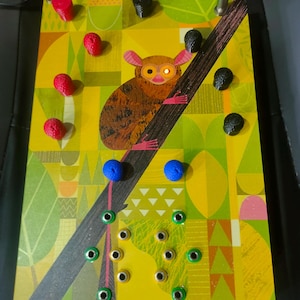 Op de afbeelding: Een rechthoekige elektronische synthesizer-module met een gele en groene geometrische achtergrond en een centrale illustratie van een spookdiertje dat aan een tak hangt. Het apparaat bevat diverse rode, zwarte en blauwe knoppen en metalen patchpunten.