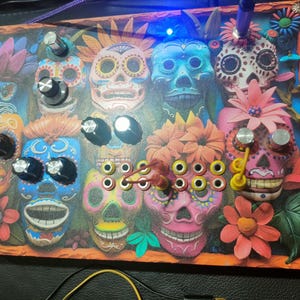Puede incluir: Panel decorativo con diseños coloridos de calaveras de azúcar del Día de Muertos. El panel incluye varias perillas y elementos decorativos, con una vibrante paleta de colores de azules, rosas, naranjas y amarillos. El panel es rectangular con un borde naranja.