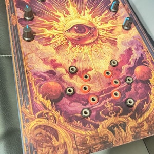 Può includere: Un dispositivo elettronico personalizzato con un'illustrazione dettagliata e colorata di un occhio all'interno di un disegno a raggiera. È dotato di più manopole e prese di ingresso, con una combinazione di colori viola e oro. Il dispositivo ha un'estetica vintage.