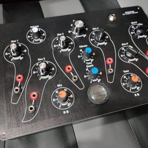 Può includere: Sintetizzatore elettronico nero con più manopole e quadranti. Dispone di controlli etichettati per volume, oscillatori, tempo e altre funzioni. Include accenti rossi e blu e un componente circolare trasparente.