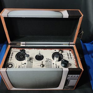 Puede incluir: Una caja abierta de estilo vintage con un diseño de televisión retro. La caja presenta un panel de control con perillas e interruptores, y un asa de cuero sintético. La combinación de colores incluye tonos de marrón, gris y negro, con una apariencia envejecida.