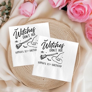 May include: Two white paper napkins with a black witch-themed design. The design includes a witch's hat, broom, spider, and the text "Witches Don't Age" and "Sophia's 30th Birthday". The napkins are on a woven tray.