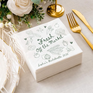 May include: A stack of white paper napkins with a green vegetable illustration and the text "Fresh Off the Market". The napkins also include the text "Sophia's Bachelorette 2025". Gold utensils are visible in the background.