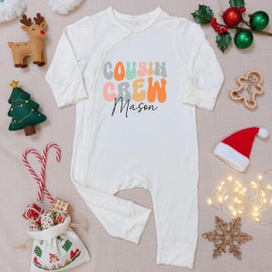 May include: Cream-colored baby onesie with the text "COUSIN CREW Mason" in colorful retro font. The onesie is surrounded by Christmas decorations, including a Santa hat, candy canes, and a felt Christmas tree.