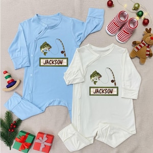 May include: Two baby rompers, one light blue and one white, each featuring a fishing-themed graphic with a fish and a fishing rod. The name "JACKSON" is printed on a wooden sign. Christmas decorations are in the background.