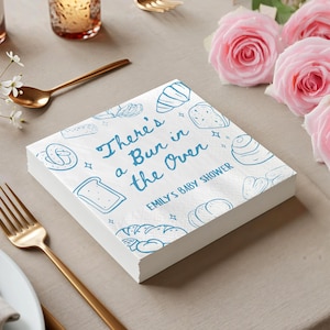 May include: A stack of white paper napkins with a blue illustrated bread design. The text "There's a Bun in the Oven" is written in blue script, with "Emily's Baby Shower" below. The napkins are on a table with gold cutlery and pink roses.