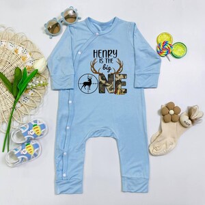 May include: Light blue baby jumpsuit with long sleeves and snap closures. The front features the text "HENRY is the big ONE" with a deer and camouflage design. Accessories include sunglasses, socks, and sandals.