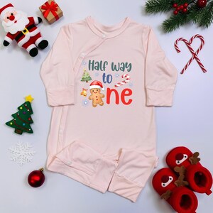 May include: A light pink baby sleeper with the text "Half way to one" and Christmas-themed graphics. The design includes a gingerbread man, Christmas tree, and candy cane. Festive holiday decor surrounds the sleeper.