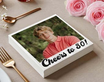 May include: A stack of white square coasters with a photo of a young person in a coral polo shirt. The text "Cheers to 30!" is printed in black script. Gold cutlery and pink roses are in the background.