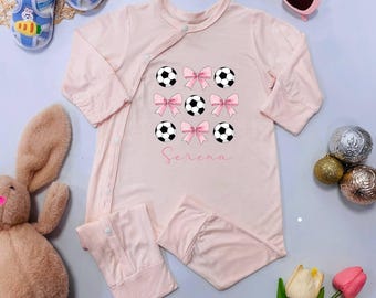 May include: A light pink baby onesie with long sleeves and snap closures. The onesie features a design of soccer balls and pink bows, with the name "Serena" printed in cursive. Additional items include baby shoes, sunglasses, and a stuffed bunny.