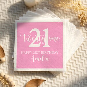 May include: A stack of white paper napkins with a pink square in the center. The pink square has the number "21" and the words "twenty one" in white script. Below the number are the words "HAPPY 21ST BIRTHDAY" and the name "Amelia".