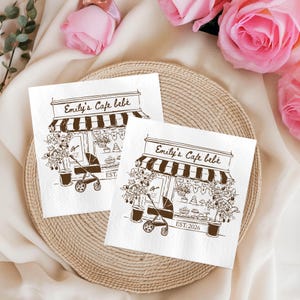 May include: Two white napkins with a brown illustration of a cafe facade, a baby stroller, and the text "Emily's Cafe bébé EST. 2026". The napkins are on a woven tray, with pink roses in the background.