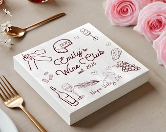 May include: A stack of white paper napkins with a burgundy wine-themed design. The napkins are printed with wine glasses, cheese, grapes, and the text "Emily's Wine Club est. 2025 Napa Valley, CA". Pink roses and gold cutlery are in the background.