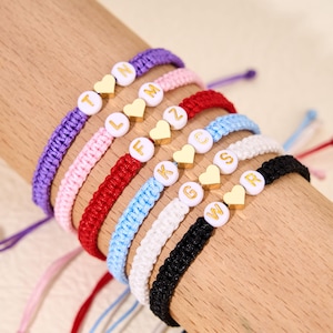 May include: A collection of personalized friendship bracelets in various colors, including purple, pink, red, blue, white, and black. Each bracelet features a letter bead and a gold heart charm. The bracelets are displayed on a wooden surface.