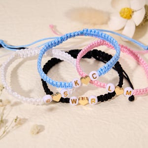May include: A collection of handmade friendship bracelets in various colors, including white, blue, black, and pink. Each bracelet features letter beads and heart-shaped charms, spelling out names or initials. The bracelets are displayed on a light-colored surface.