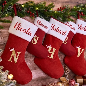 Peut inclure: Cinq bas de Noël rouges avec des revers en fausse fourrure blanche sont suspendus sur un fond en bois. Chaque bas est personnalisé avec un nom brodé en rouge et une initiale dorée. Décor de vacances festif.