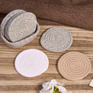 May include: A set of woven coasters in various colors, including white, beige, and gray, with a matching holder. The coasters are arranged on a wooden surface, showcasing their spiral design and texture. A decorative floral arrangement is also present.