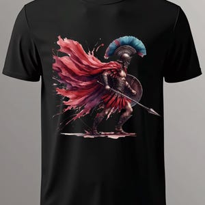 May include: Black t-shirt featuring a watercolor illustration of a Spartan warrior. The warrior is depicted in armor, holding a spear and shield, with a red cape and a blue and red plumed helmet. The design has a dynamic, artistic style.